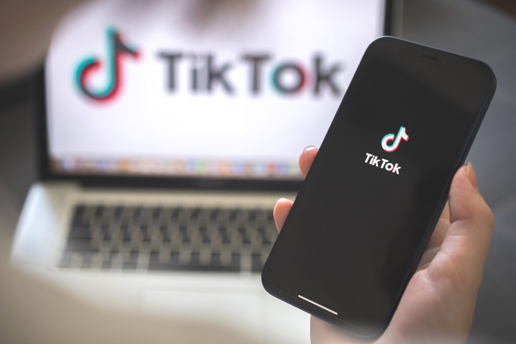 Een persoon houdt een smartphone vast met het TikTok-logo erop, terwijl op de achtergrond een laptop te zien is met het TikTok-logo op het scherm